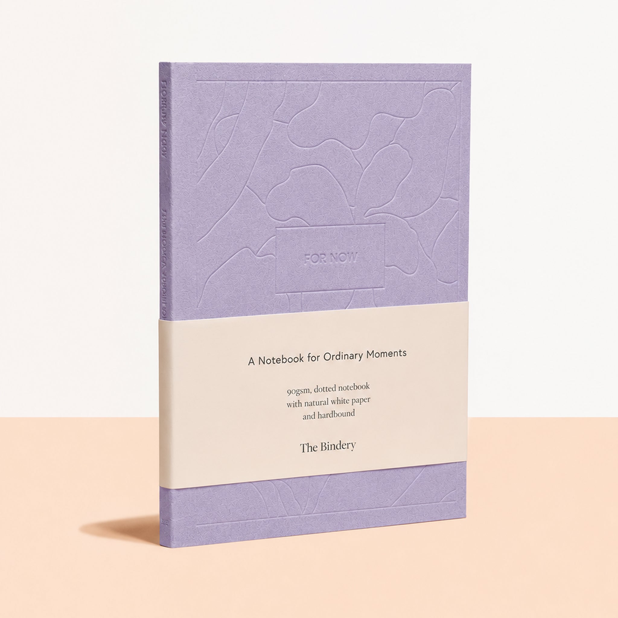 A lavender notebook with a cream paper band around the middle, standing upright on a pale peach surface against a light beige background. The front cover has an embossed abstract pattern, and the band reads 'A Notebook for Ordinary Moments' with 'The Bindery' at the bottom.