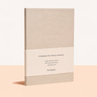 A notebook with a textured beige cover and a cream paper band around the middle, standing upright on a peach-colored surface against a white background. The band reads 'A Notebook for Ordinary Moments' and shows the brand 'The Binder'.