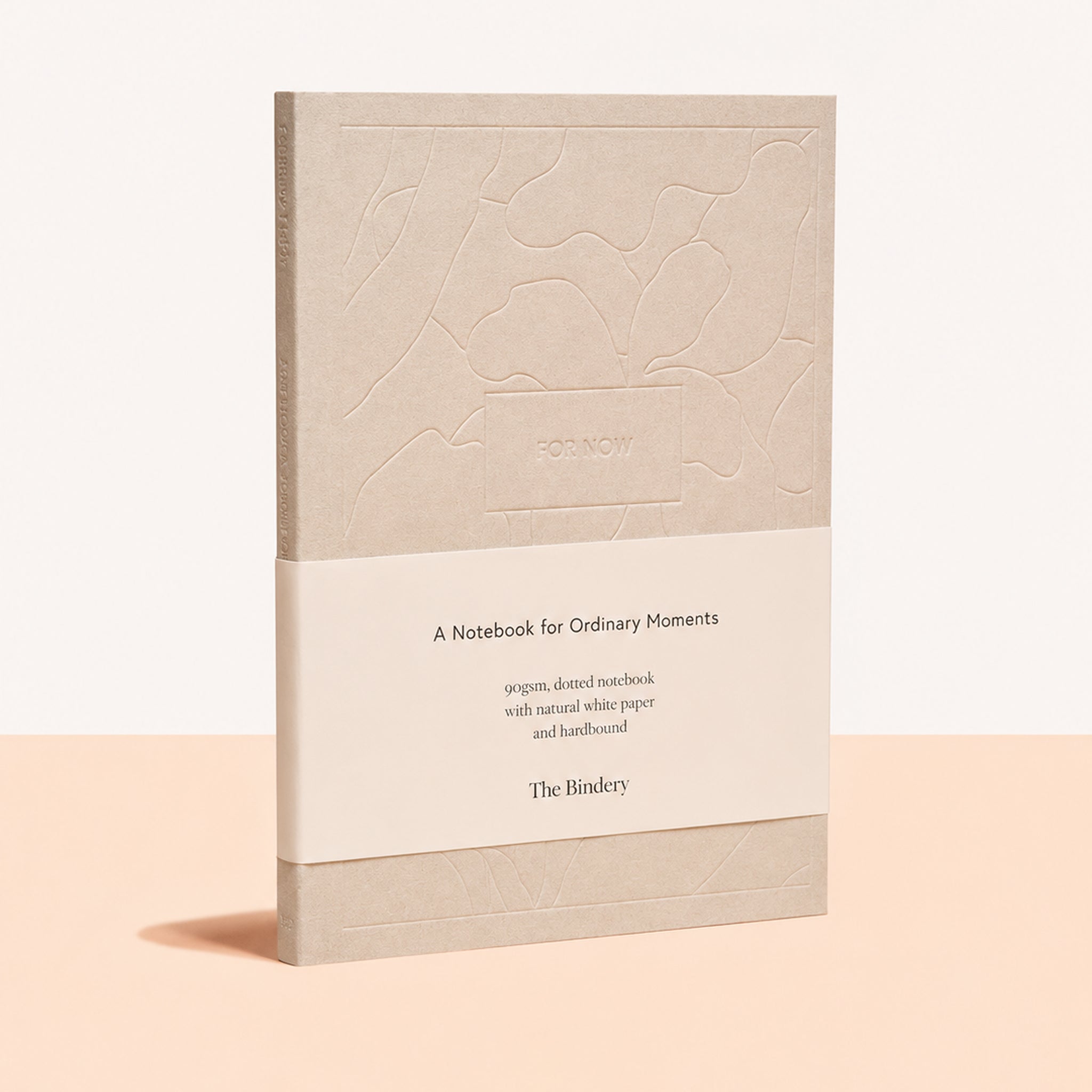 A notebook with a textured beige cover and a cream paper band around the middle, standing upright on a peach-colored surface against a white background. The band reads 'A Notebook for Ordinary Moments' and shows the brand 'The Binder'.